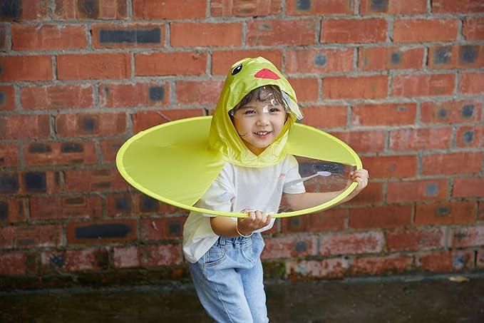 Smiling child wearing yellow kid's duck rain cover with hood and clear visor standing by brick wall